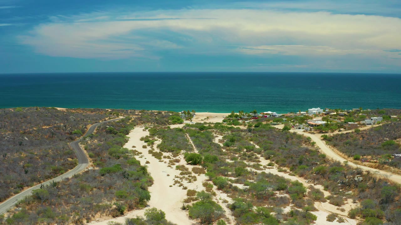 High coastal view from San Jose Del Cabo, Mexico, showing scrub-covered terrain, a pale sandy wash through the center, scattered greenery and buildings by the beach, open ocean, soft clouds, and no visible people.