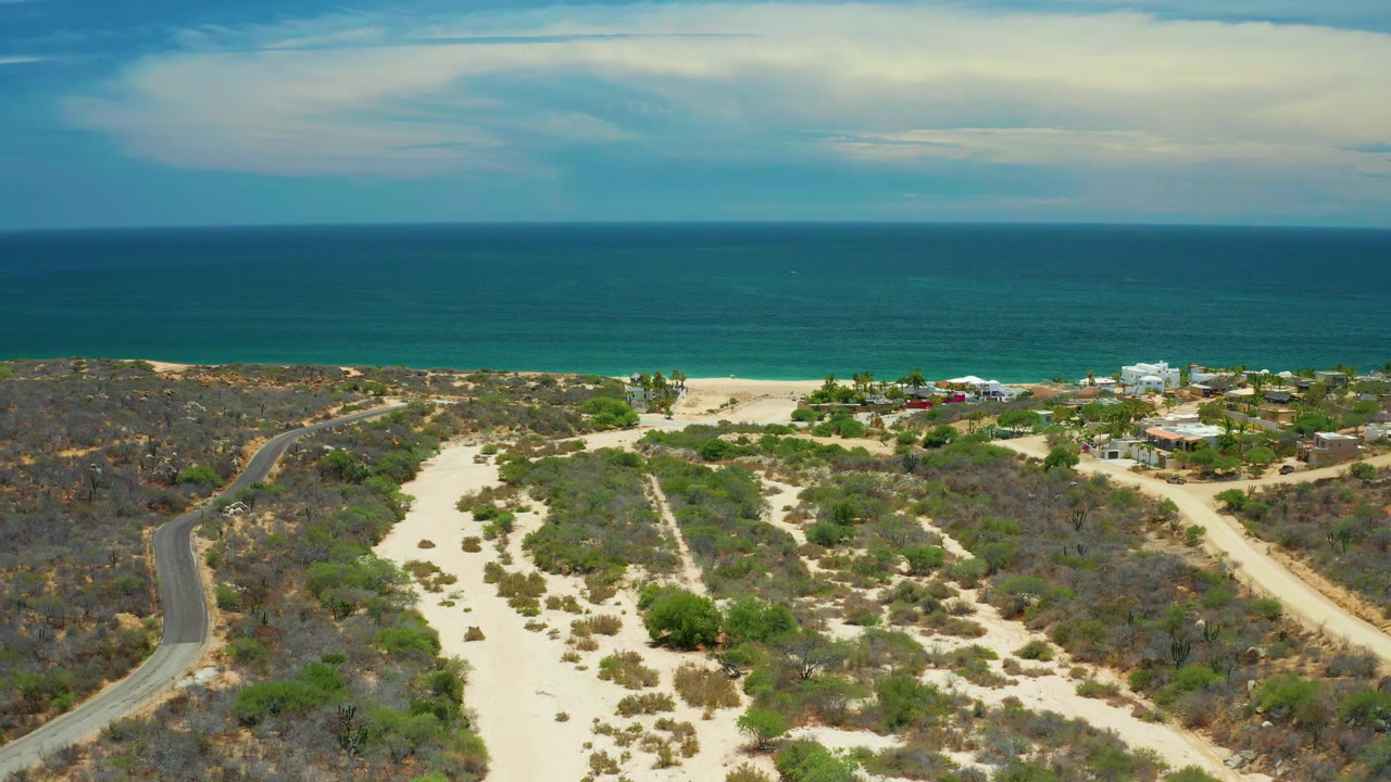 Drone-like coastal view with sandy land, brush, beach structures, and ocean.