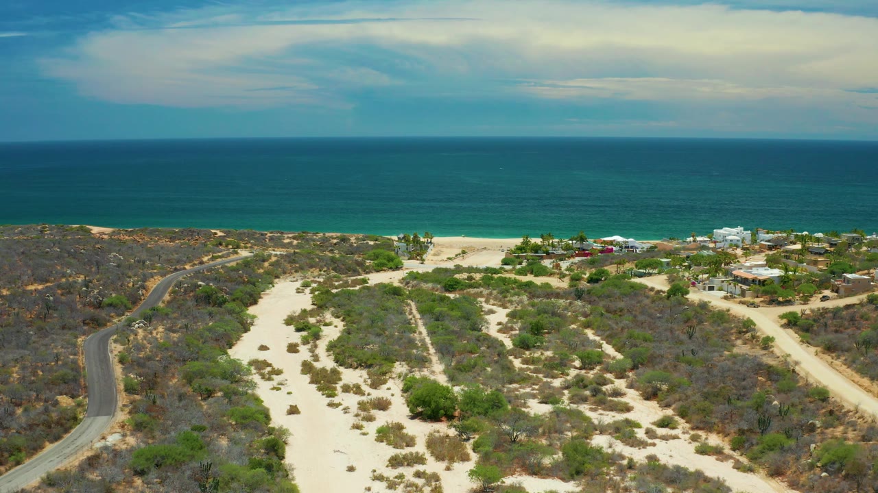 Aerial San Jose Del Cabo frame showing sandy open terrain and scrub in the foreground, the road along the left side, beachfront buildings to the right, a calm ocean horizon, and no visible people.