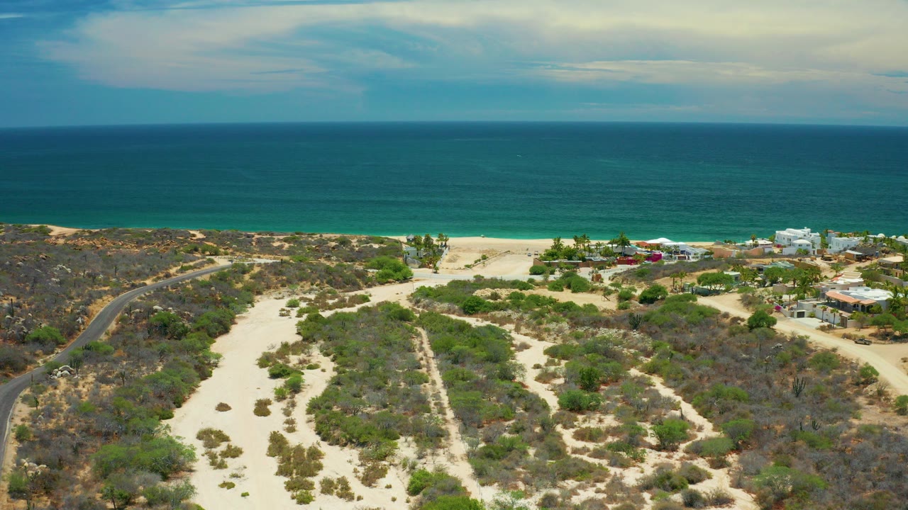 Aerial frame over San Jose Del Cabo desert scrub and pale sand paths leading toward a beach and turquoise ocean, with no visible people.