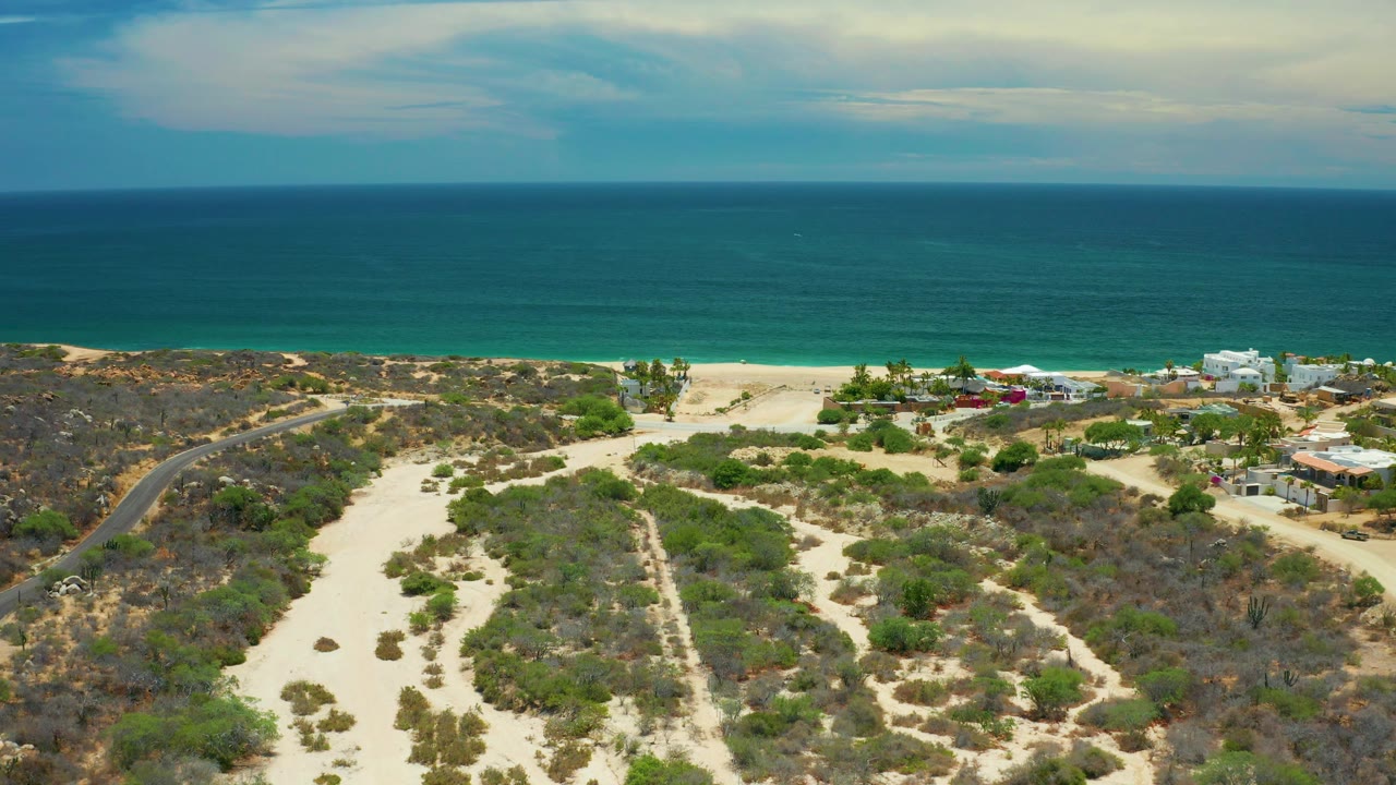 Aerial view in San Jose Del Cabo with a curved road through scrubland, scattered coastal buildings near the beach, ocean water beyond, and no visible people.
