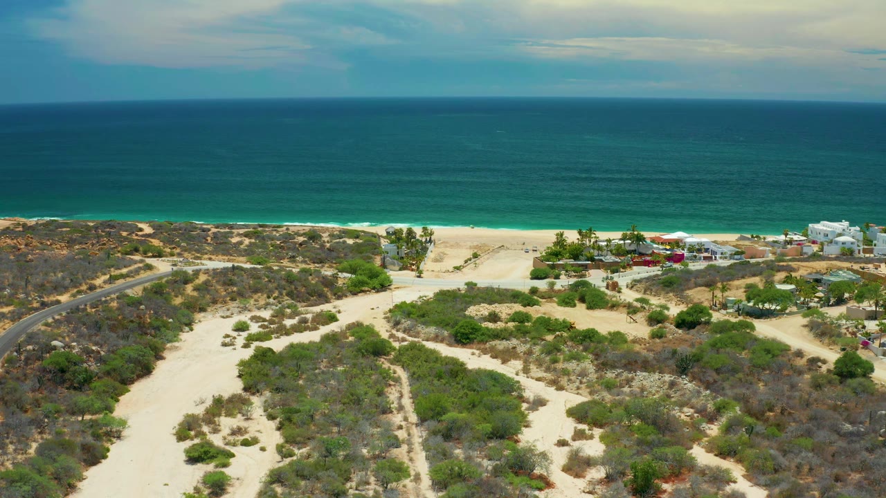 Second aerial frame in San Jose Del Cabo showing a broad green-blue ocean band, thin surf, beach, dune scrub, a road at left, shoreline buildings at right, and no visible people.