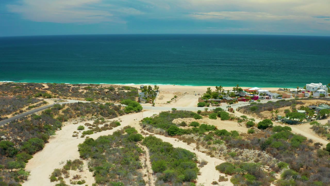 Aerial coastline view with ocean, sandy beach, road, scrub, and buildings.