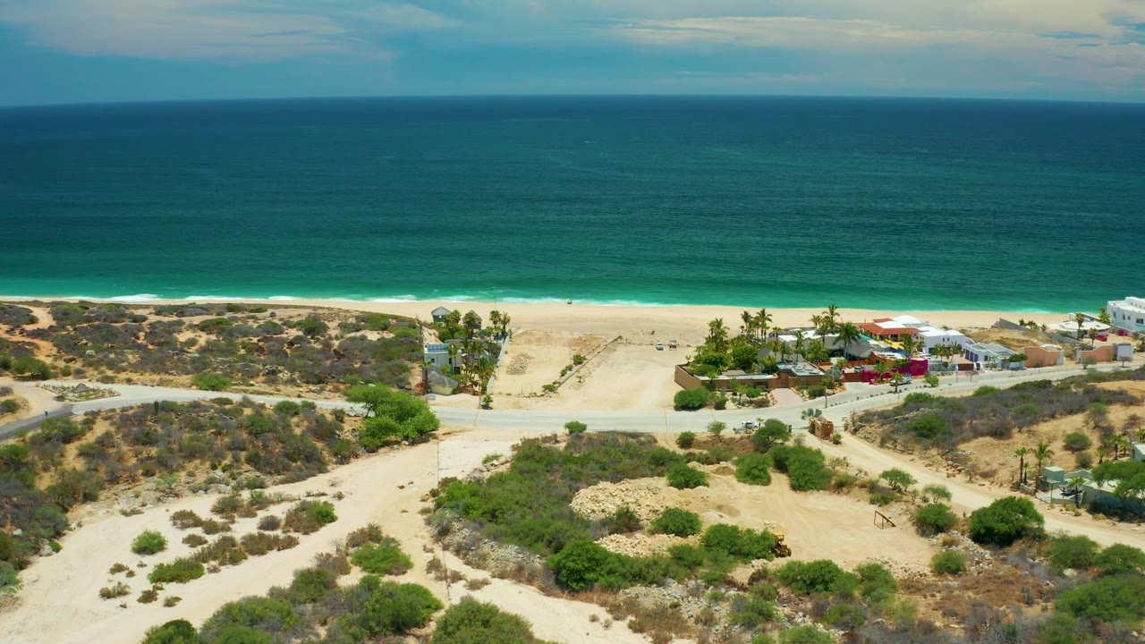 Elevated San Jose Del Cabo frame with scrub-covered dunes in front, a row of small homes near the sand, blue-green sea beyond, and no visible people.