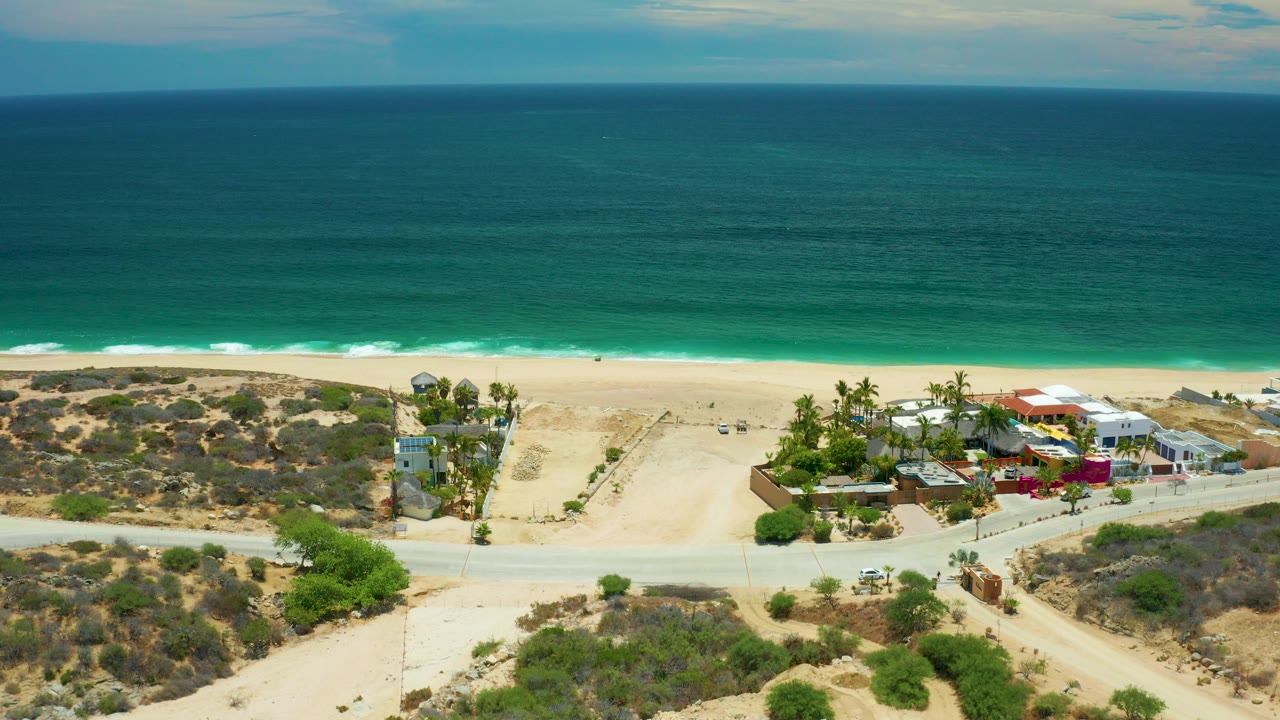 Aerial frame over San Jose Del Cabo showing turquoise ocean, a pale beach, scrubby dunes, a coastal road, palm trees, and low beachfront buildings with no visible people.