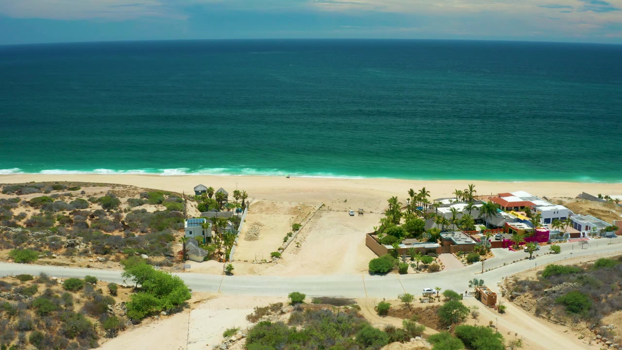 San Jose Del Cabo aerial frame showing a wide band of turquoise sea, bright sand, dry dunes, a road junction, palm trees, and colorful low buildings, with no visible people.
