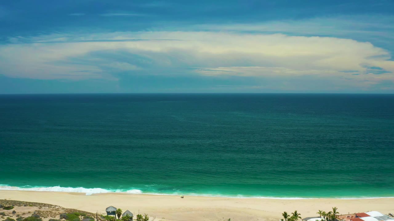 San Jose del Cabo shoreline view with teal water, a straight horizon, pale cloud bands, a thin beach strip, and no visible people.