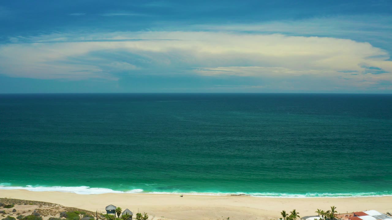Wide elevated view of a San Jose Del Cabo beach with teal ocean, pale sand, white surf, broad cloud cover, and no visible people at 0:00.