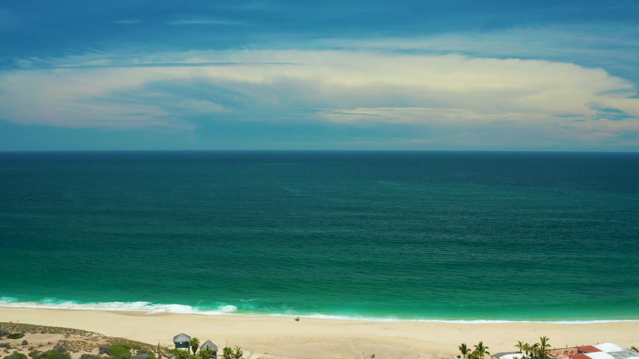 San Jose Del Cabo, Mexico beach from a high wide view, with green ocean water, a pale sandy shoreline, cloud cover, and no visible people.
