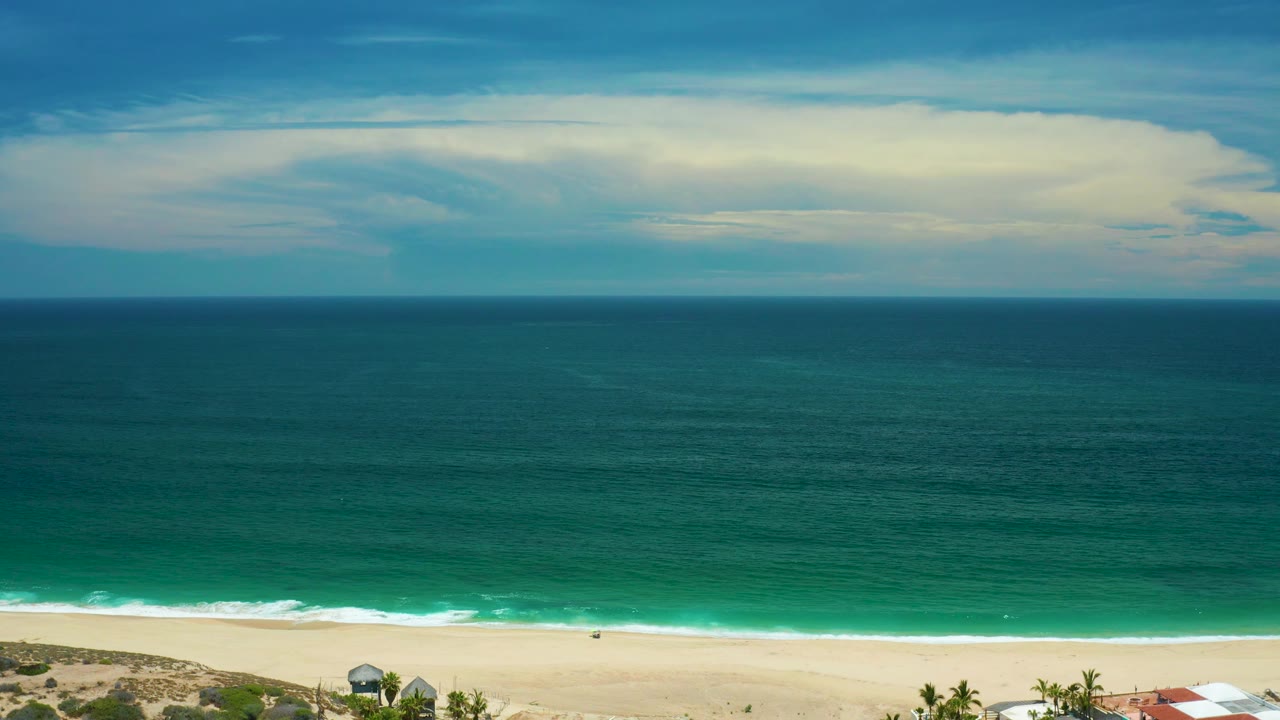 A wide San Jose Del Cabo coastal frame shows a straight horizon, muted cloud light, green ocean bands, pale beach, and no visible people.