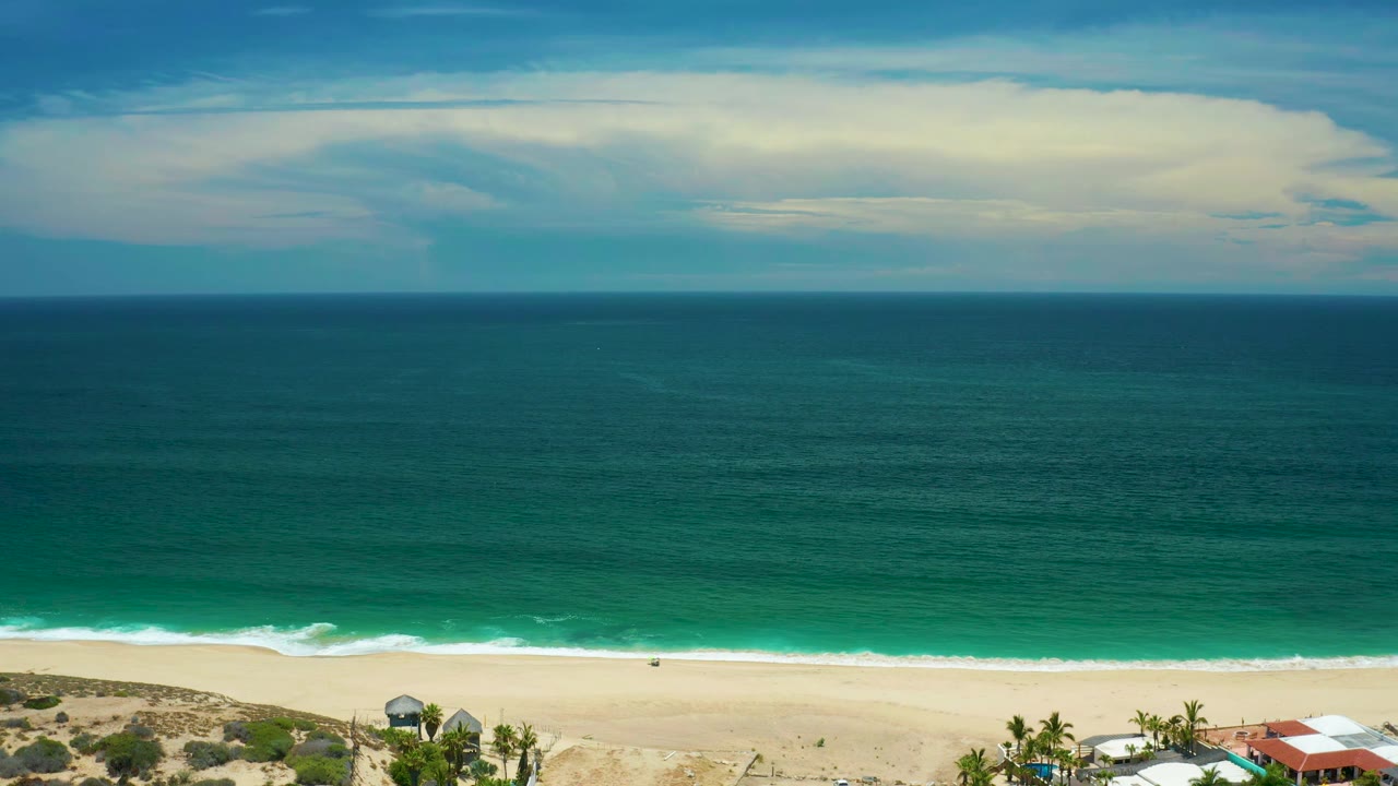 Aerial frame over San Jose Del Cabo with the ocean filling most of the view, a thin white surf band, pale sand, and partial beachfront roofs along the lower right, with no visible people.