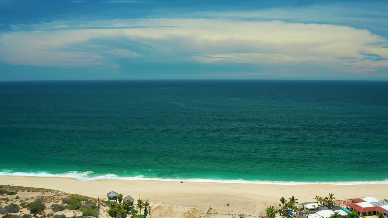 San Jose Del Cabo beach view with darker blue-green water farther out, brighter water near the surf, a narrow beach strip, and small structures cropped at the lower right, with no visible people.