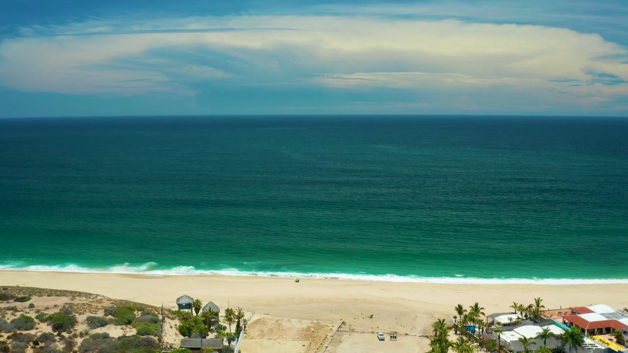 Aerial view over a San Jose del Cabo beach with pale sand, green-blue ocean bands, a level horizon, layered cloud, and no visible people.