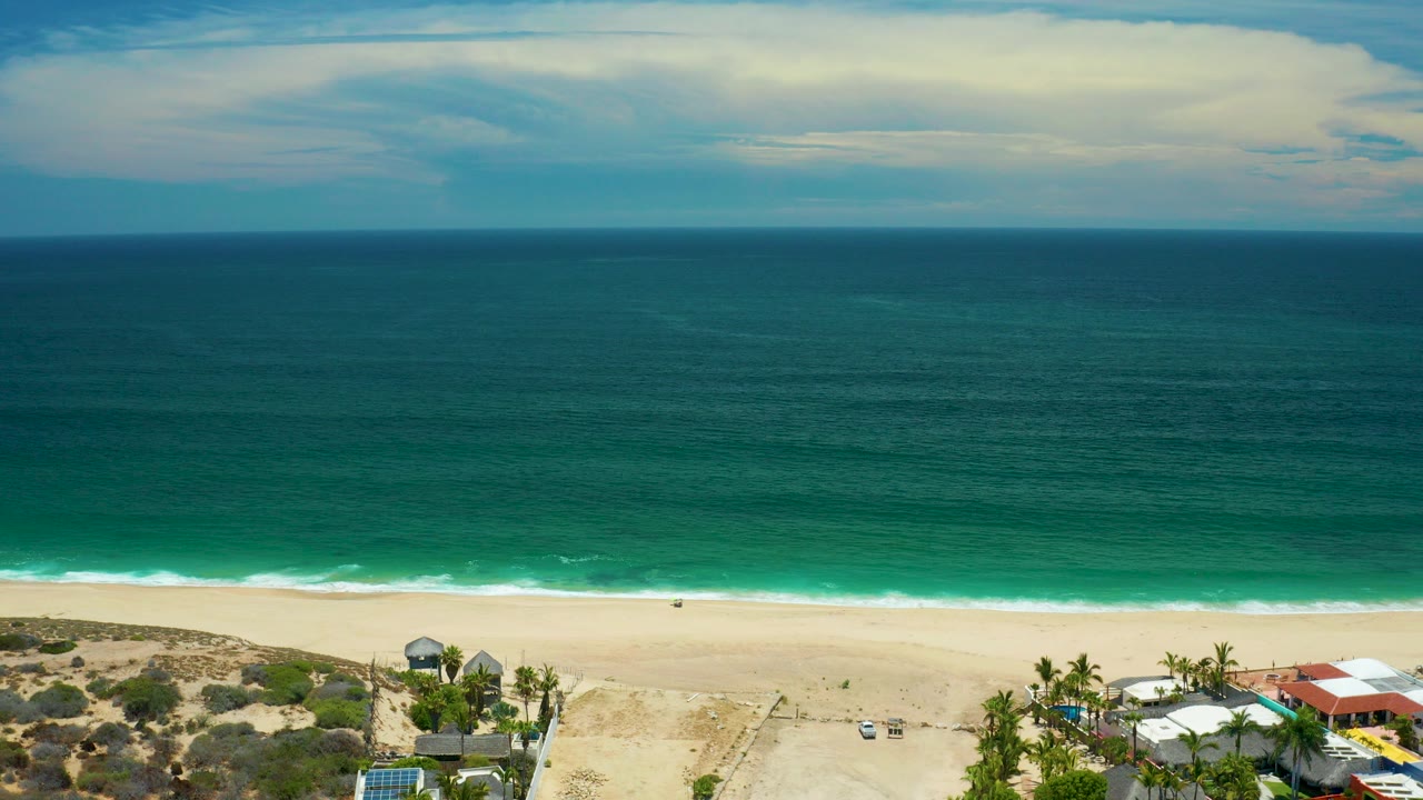 Wide aerial view of the San Jose del Cabo coast with teal ocean across most of the frame, pale sand at the bottom, palms and low buildings near the shore, and no visible people.