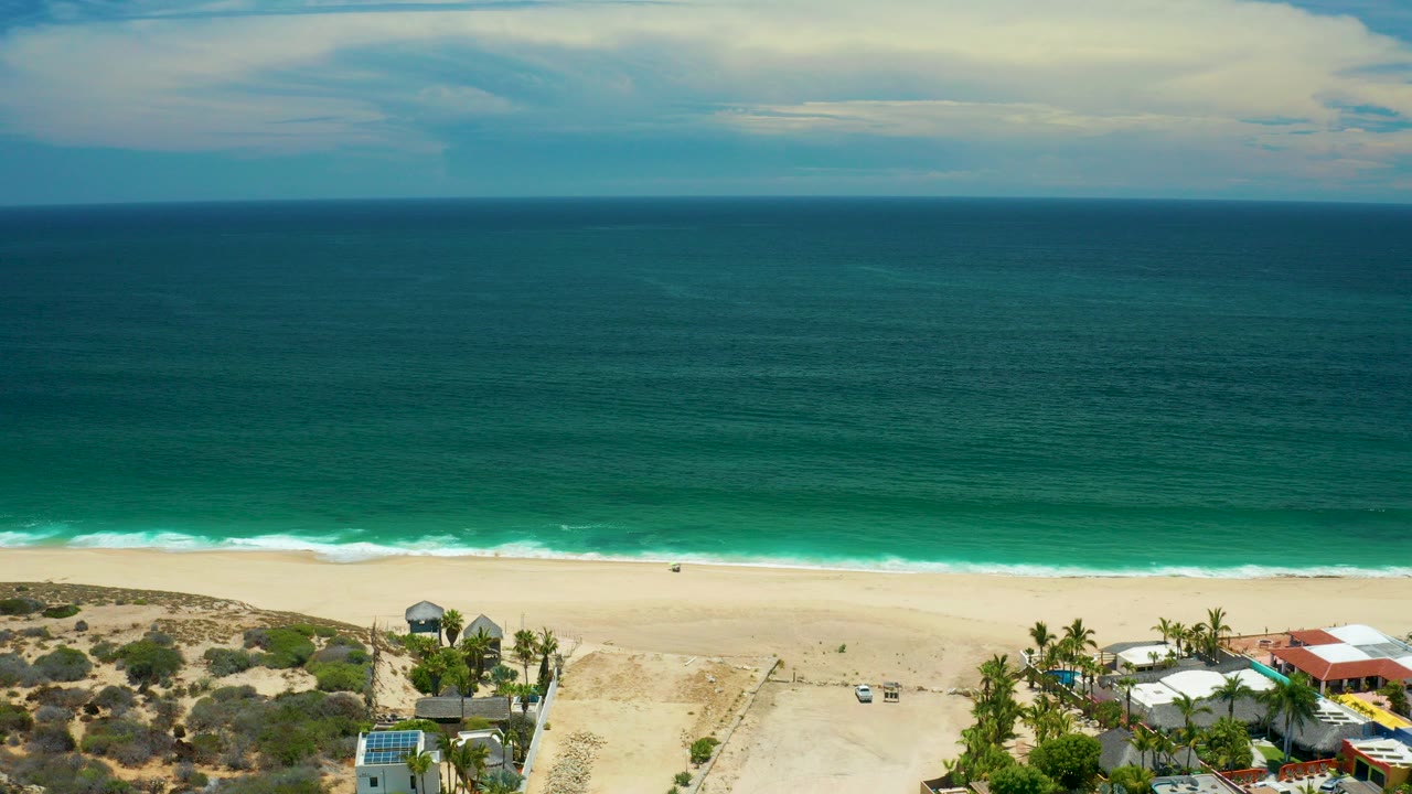 Opening elevated view over San Jose Del Cabo coastline with teal ocean, pale sand, scrub dunes, palm trees, partial beachfront roofs, and no visible people.