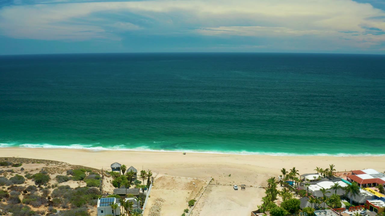 Wide San Jose Del Cabo shoreline view with deep teal water, bright surf at the beach edge, sandy open ground, palms, a red roof, and no visible people.