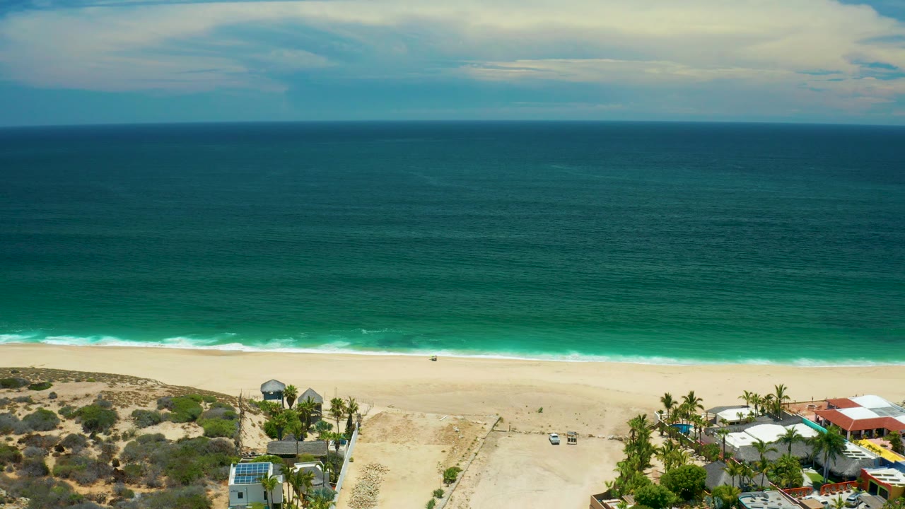 Ocean and beach from above with clouds, surf, sand, dunes, palms, and low buildings.