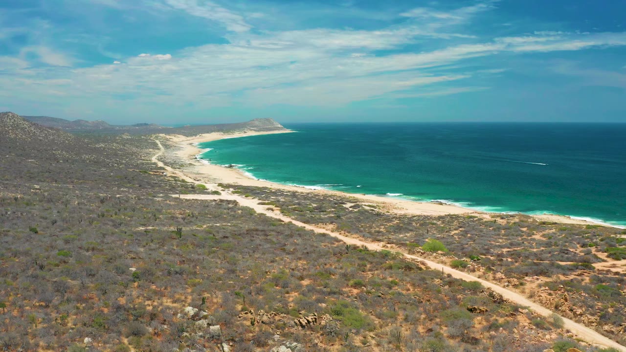 Opening aerial view over San Jose Del Cabo, Mexico, with dry scrubland leading to a curving sandy beach, blue sea, distant hills, and no visible people.