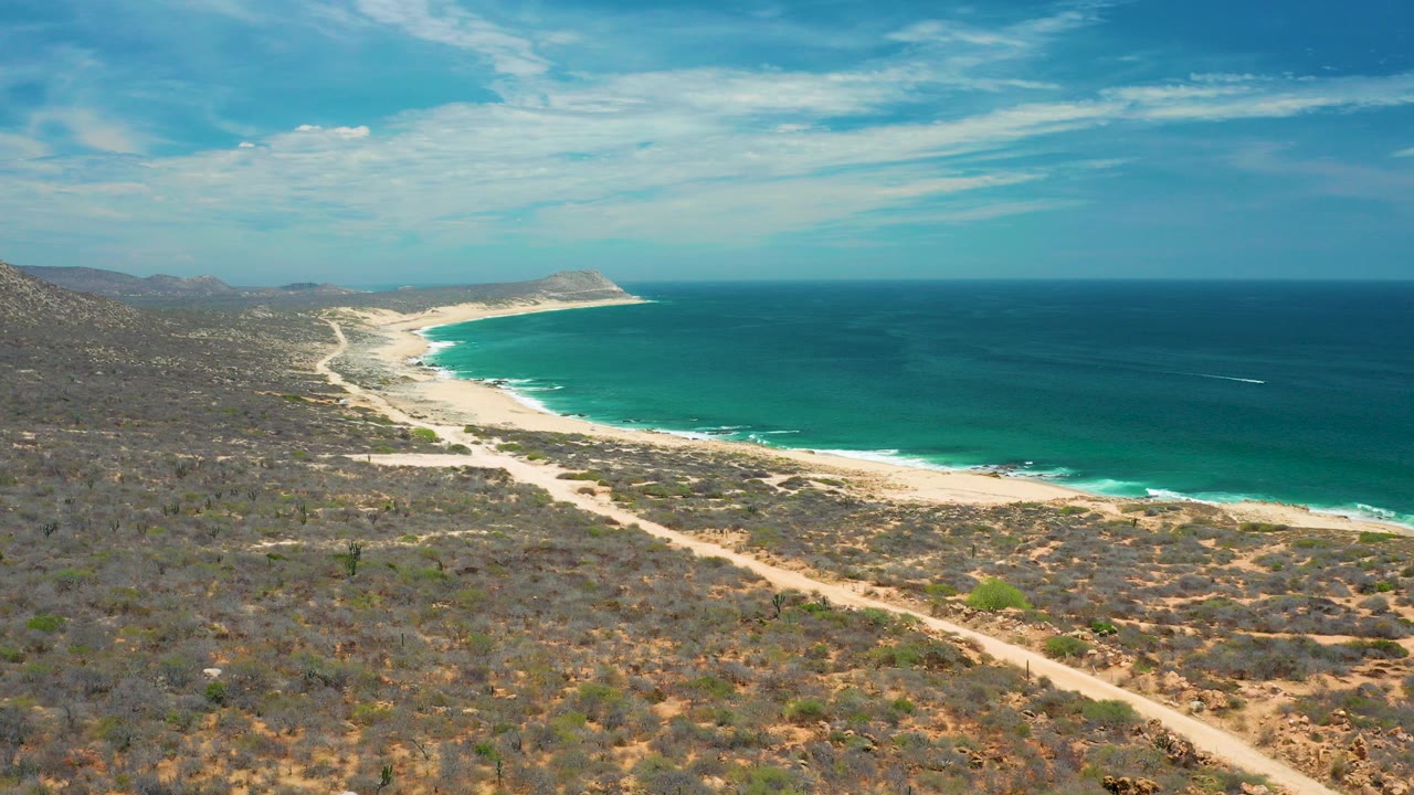 Aerial view in San Jose Del Cabo, Mexico, looking over dry coastal scrub and a sandy track toward a curving white beach, turquoise surf, deep blue ocean, distant headland, and no visible people.