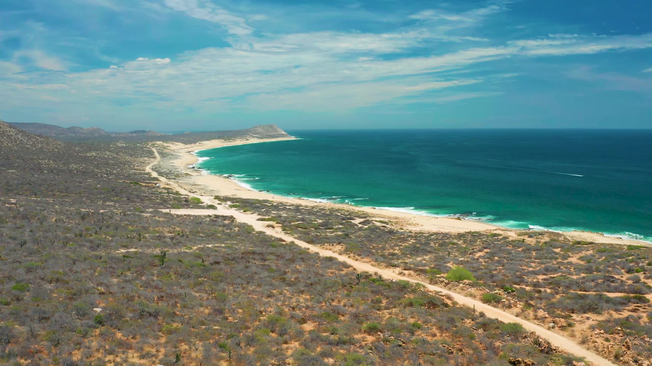 High coastal view with diagonal sandy road, bright beach, surf line, and open ocean.