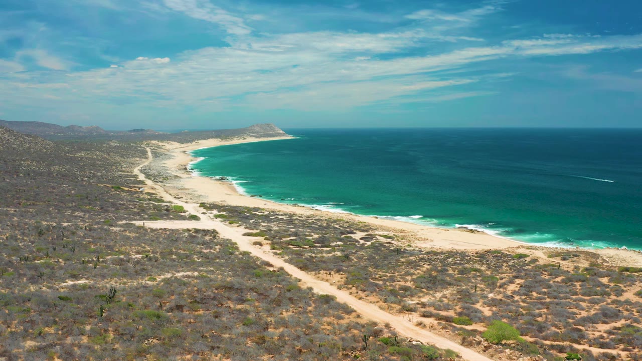 High aerial view in San Jose Del Cabo with the beach running diagonally through the frame, teal water to the right, desert brush to the left, and no visible people.