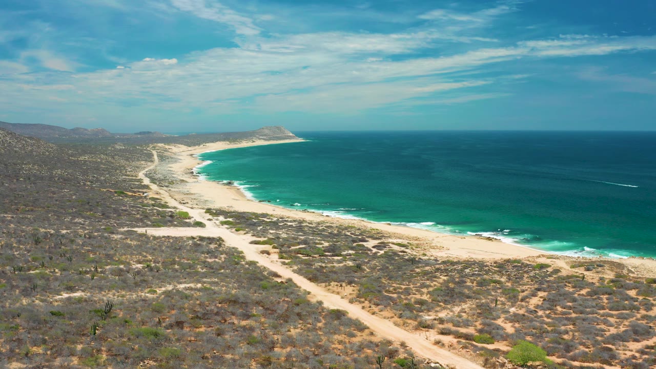 Wide San Jose Del Cabo coastal frame with an empty pale beach, layered blue ocean, scrub-covered land, low hills, cloud streaks, and no visible people.