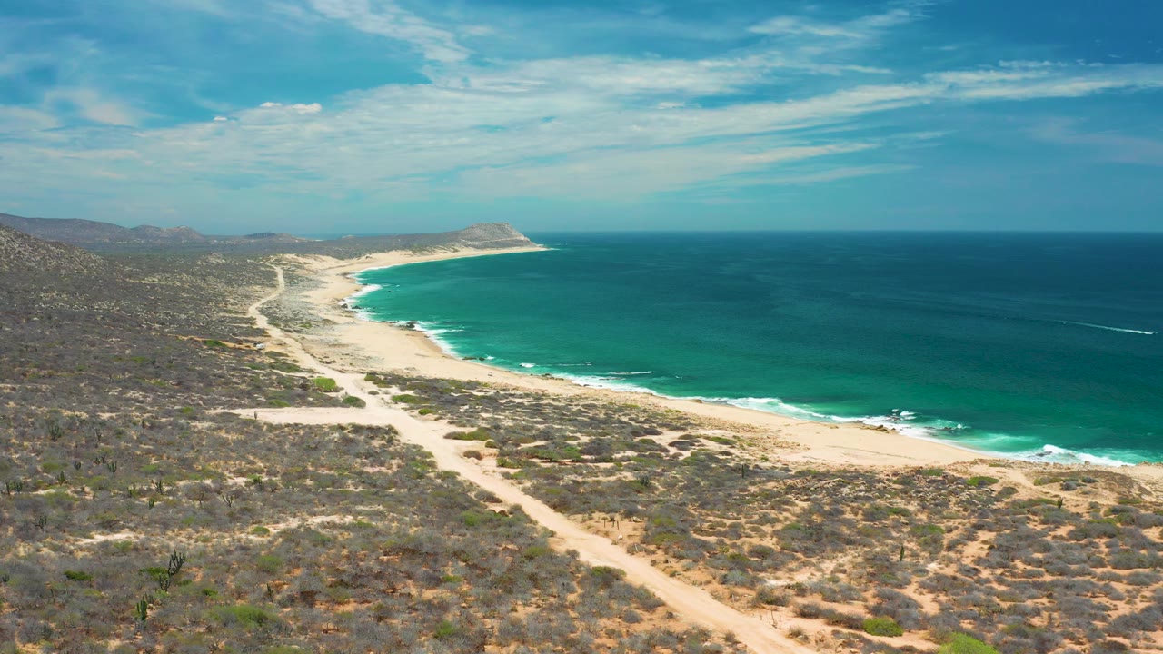 Aerial view over San Jose Del Cabo with scrubland and dirt paths in front, a bright surf edge along the beach, open water beyond, and no visible people.