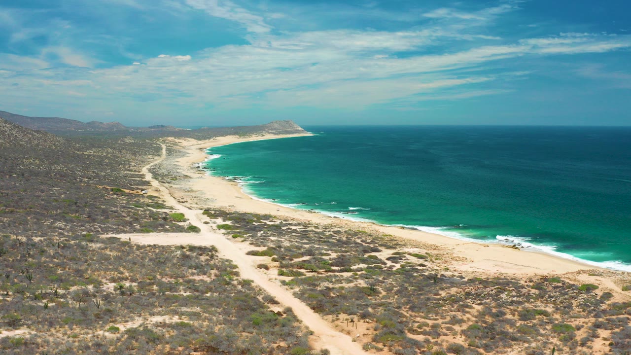 Opening aerial frame over the San Jose Del Cabo coast, with arid scrub in the foreground, a pale sandy beach curving beside blue water, and no visible people.
