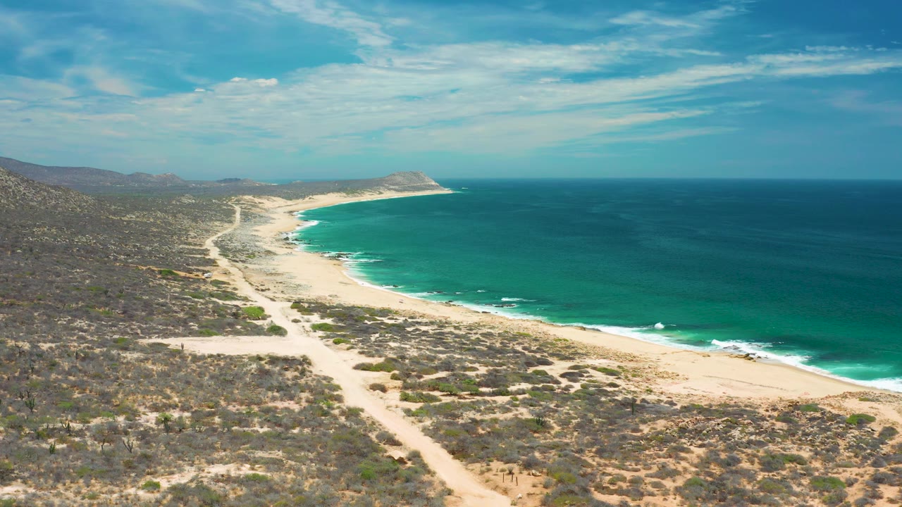 San Jose Del Cabo coastal aerial with scrub-covered terrain in the foreground, a pale beach cutting through the center, open ocean to the right, and no visible people.