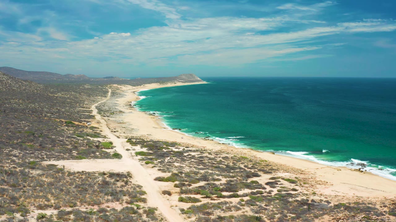 Wide aerial frame of San Jose Del Cabo beach where white waves trace the pale shoreline and scrubland fills the inland side, with no visible people.