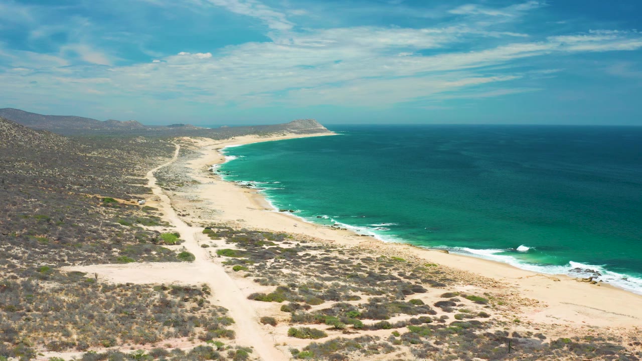 Aerial view of the curving San Jose Del Cabo shoreline with deep blue water offshore, green-blue shallows near sand, rocky scrub, and no visible people.