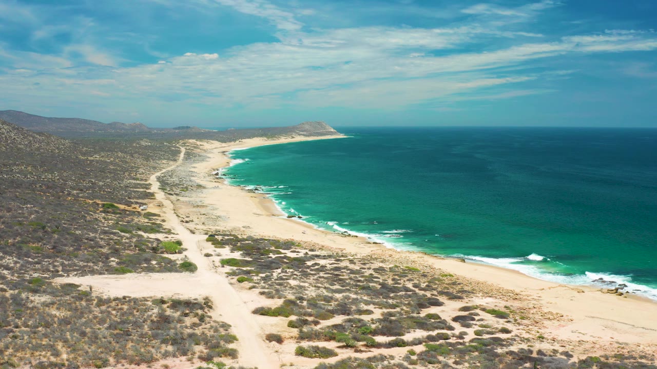 High coastal view in San Jose Del Cabo with scrub-covered foreground, pale sand, small breaking waves, broad ocean, and no visible people.
