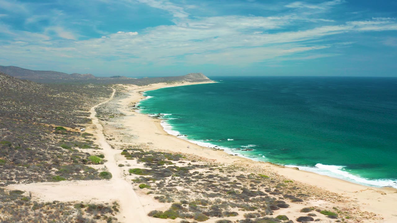 Aerial view of a sandy coast, blue-green sea, scrubland, and distant hills.