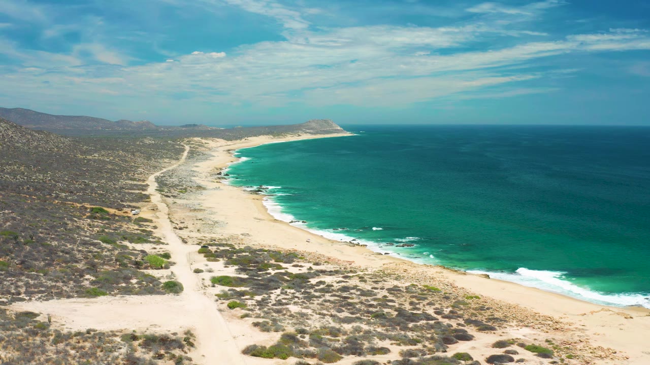 Early aerial view in San Jose Del Cabo, Mexico with the shoreline running diagonally through the frame, surf bands along the beach, scrubland at left, open water at right, and no visible people.