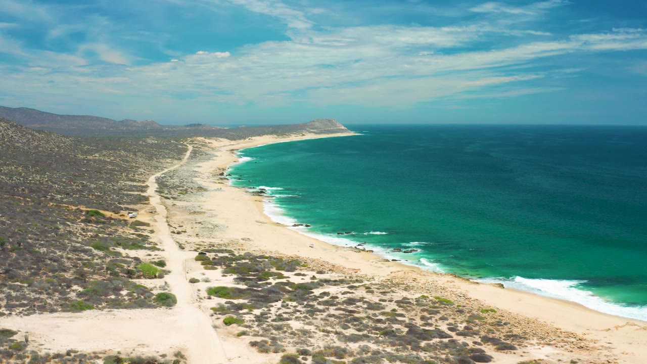 Mid-clip aerial frame in San Jose Del Cabo, Mexico with scrub and sand tracks in the foreground, a curving beach through the middle, teal surf at the edge, distant hills, and no visible people.