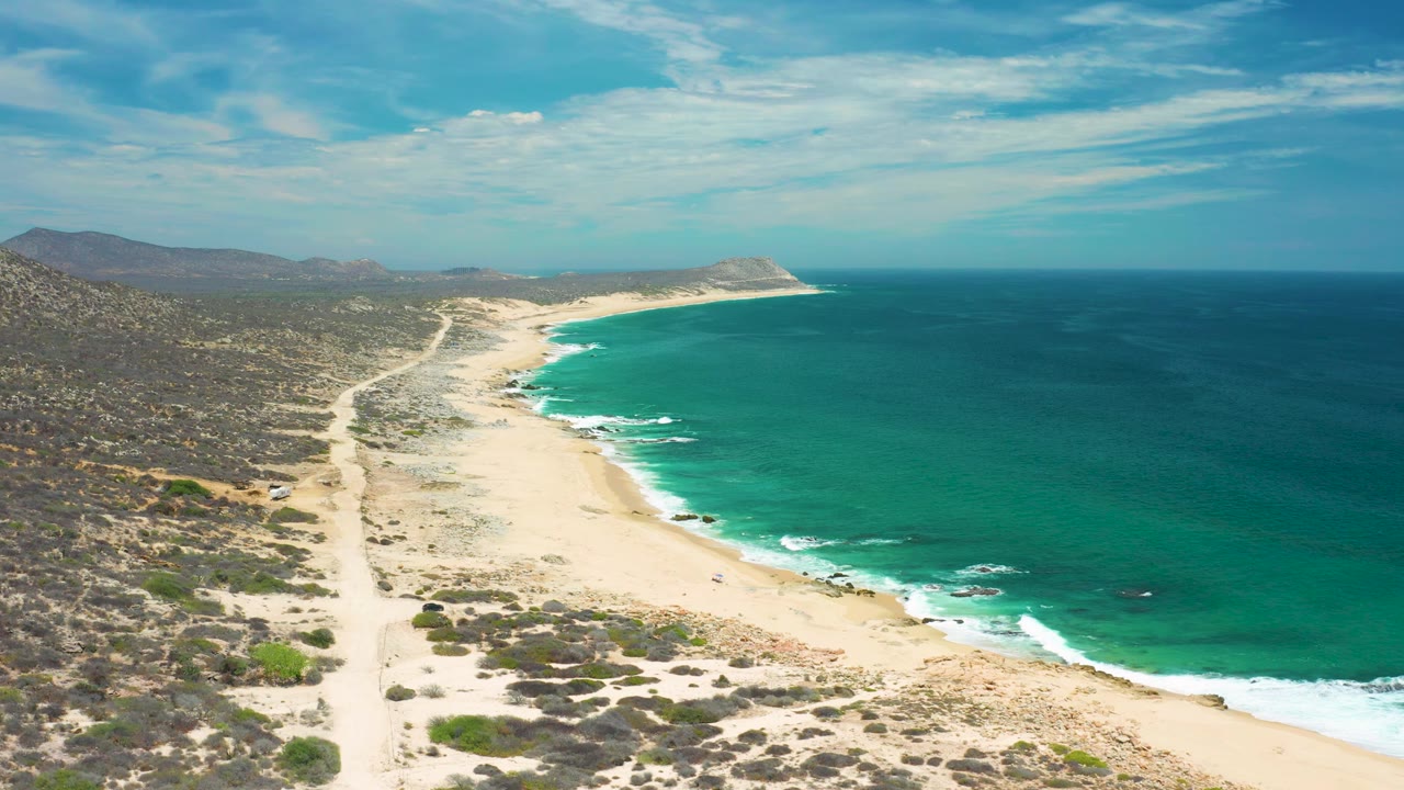 Drone-height frame in San Jose del Cabo, Mexico showing bright sand, small surf lines, a winding dirt road through scrub, low mountains on the left, open ocean on the right, and no visible people.