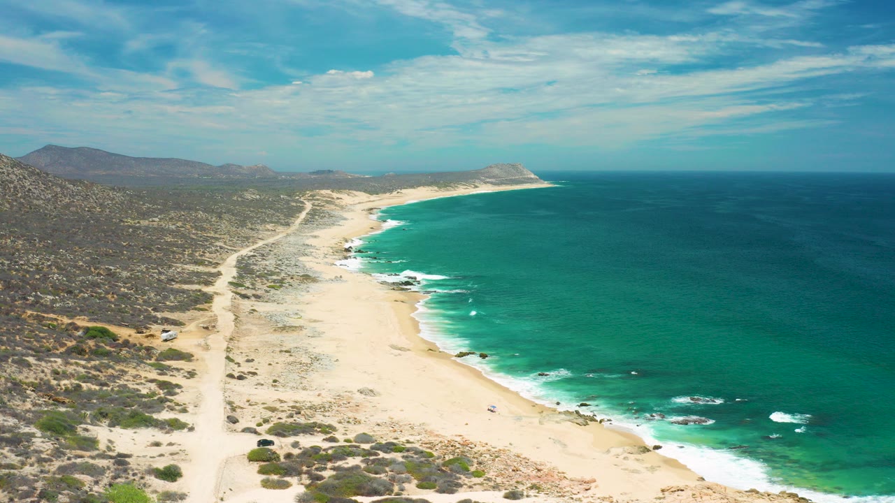 Aerial frame over a San Jose del Cabo beach with pale sand curving along green sea, scrubland on the left, distant hills, scattered clouds, and no visible people.
