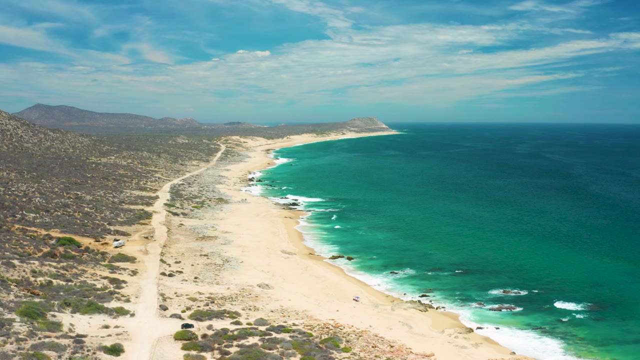 Drone-like frame above a San Jose del Cabo beach, showing empty sand, green water with white surf streaks, rocky patches near shore, inland scrub, hills, and no visible people.