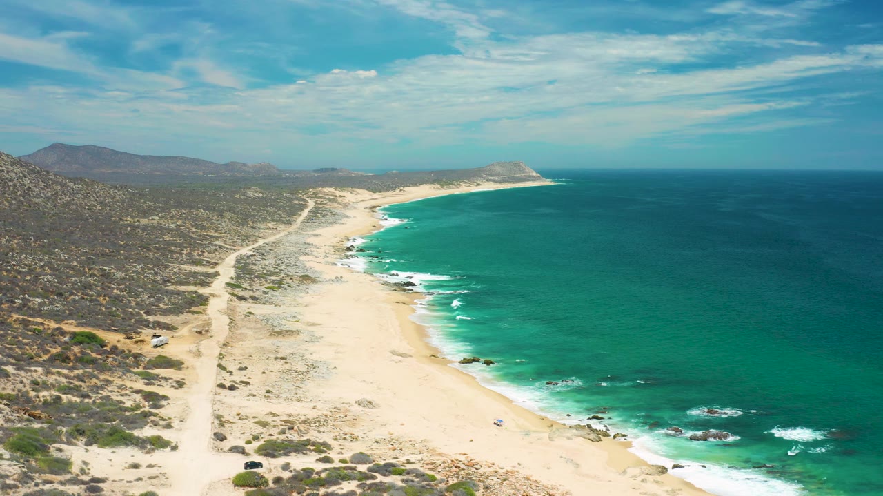 Aerial view along the San Jose del Cabo coast with a dirt track crossing scrubland, a pale beach below, green sea to the right, low white surf, distant hills, and no visible people.