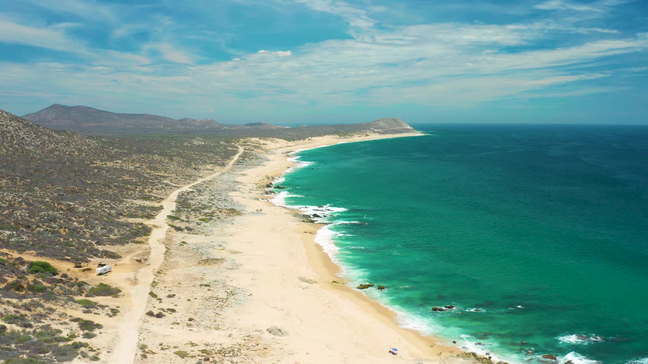 Aerial view of a San Jose Del Cabo beach where pale sand curves along turquoise water, with scrubland and a dirt track on the left and distant hills under a bright sky.