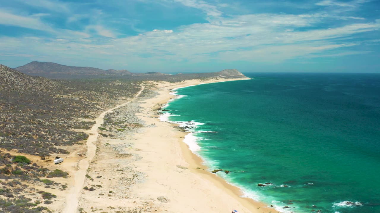 San Jose Del Cabo beach seen from above, with blue-green water filling the right side, pale sand in the center, scrubby terrain on the left, and mountains in the distance.