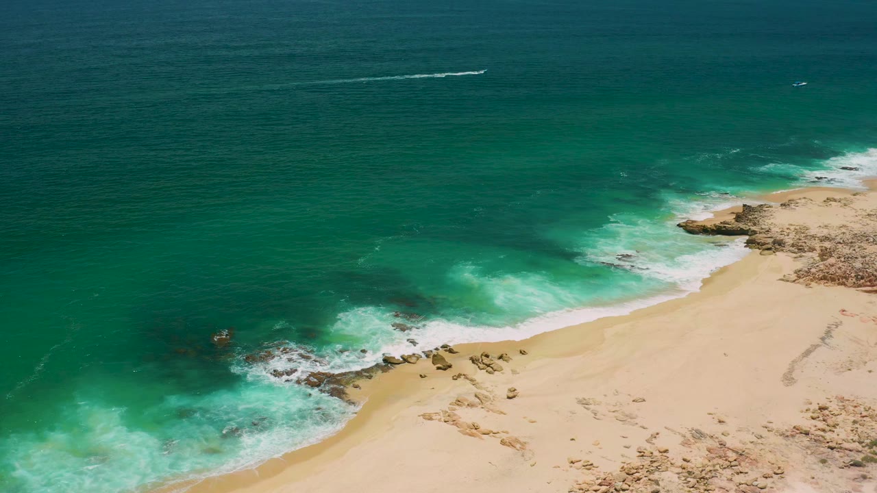 Aerial view in San Jose Del Cabo, Mexico with turquoise water spreading toward pale sand, clustered rocks near the surf line, dry terrain at right, and no visible people.