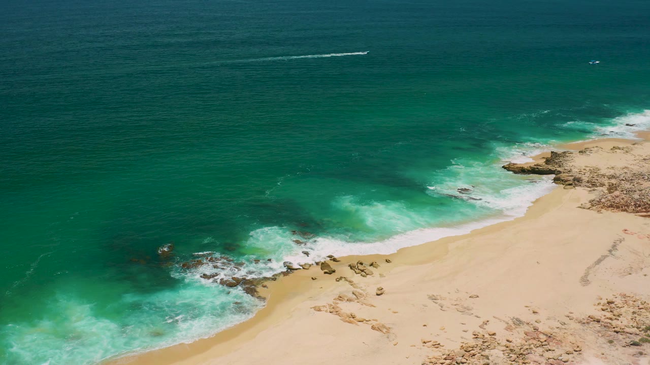 San Jose Del Cabo aerial frame showing a broad field of green water above a diagonal sandy shoreline, white foam at the rocks, a faint offshore craft or wake, and no visible people.