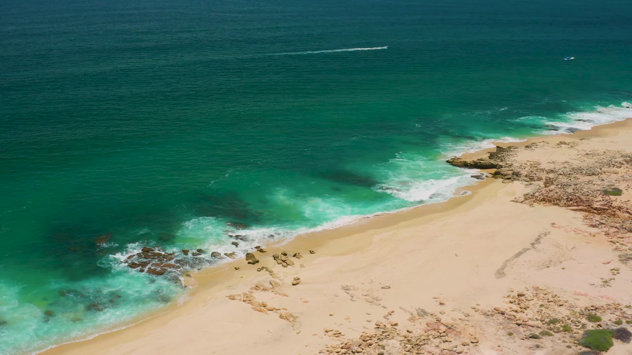 Drone-style view over San Jose Del Cabo coastline with white waves curling onto a pale beach, rocks at the waterline, and no visible people.