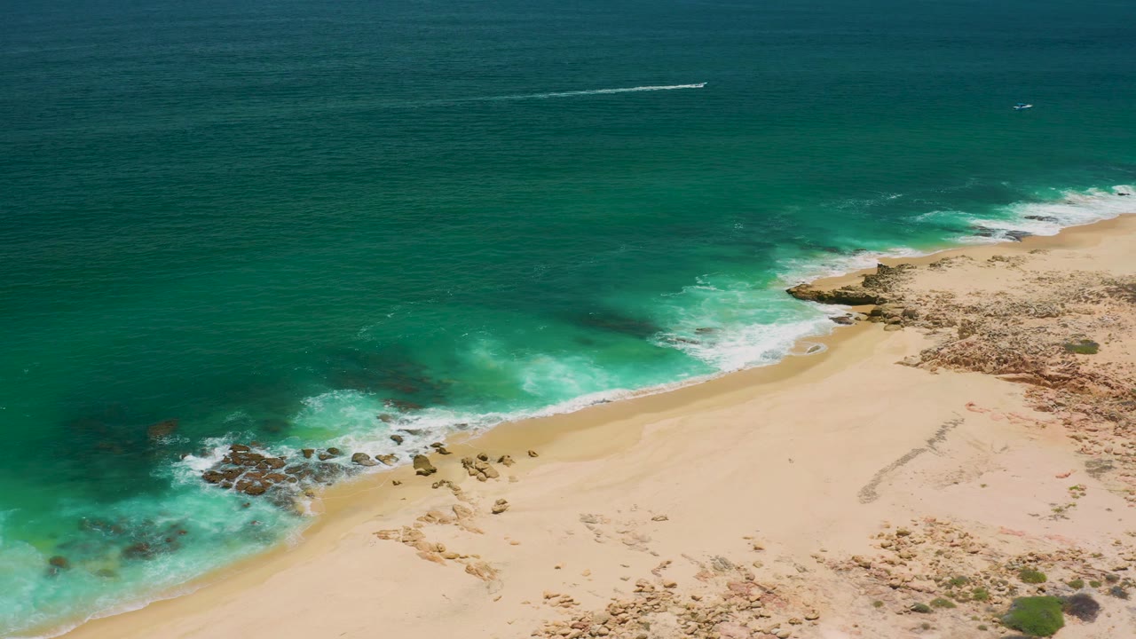 High aerial view in San Jose Del Cabo, Mexico with blue-green water washing around dark rocks beside a broad sandy beach and no visible people.