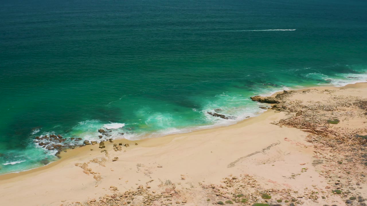 San Jose del Cabo aerial frame showing a broad band of ocean, foamy surf near rocks, pale beach, and no visible people.