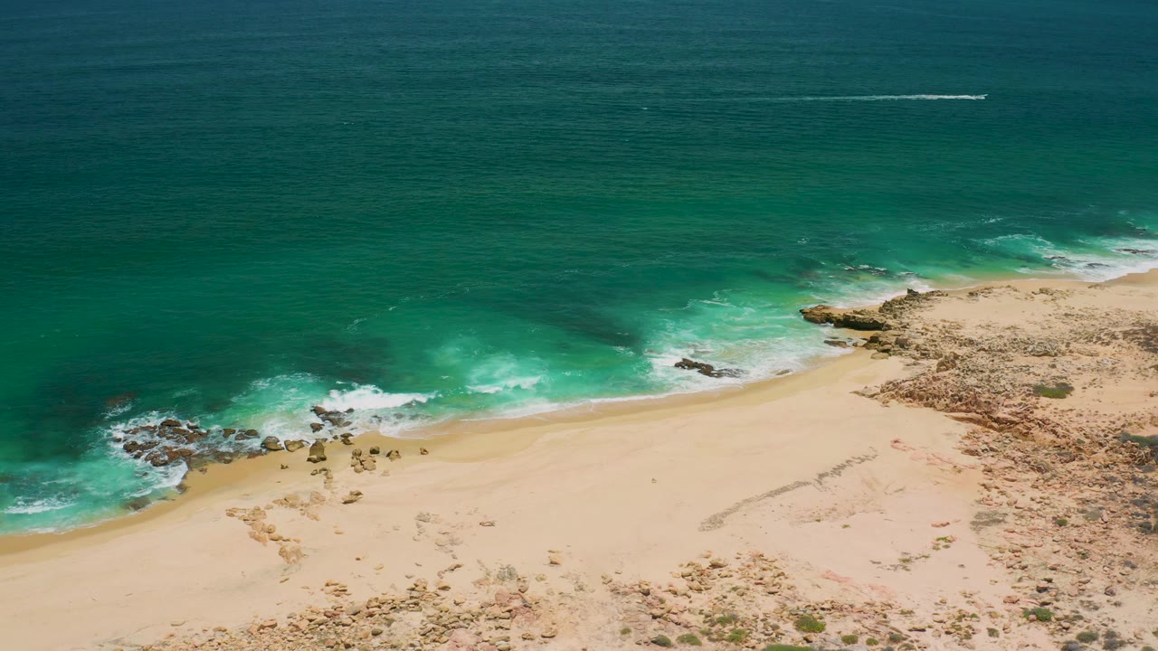 Aerial frame over San Jose del Cabo with surf spreading along the sand and rocky shoreline, and no visible people in the beach area.