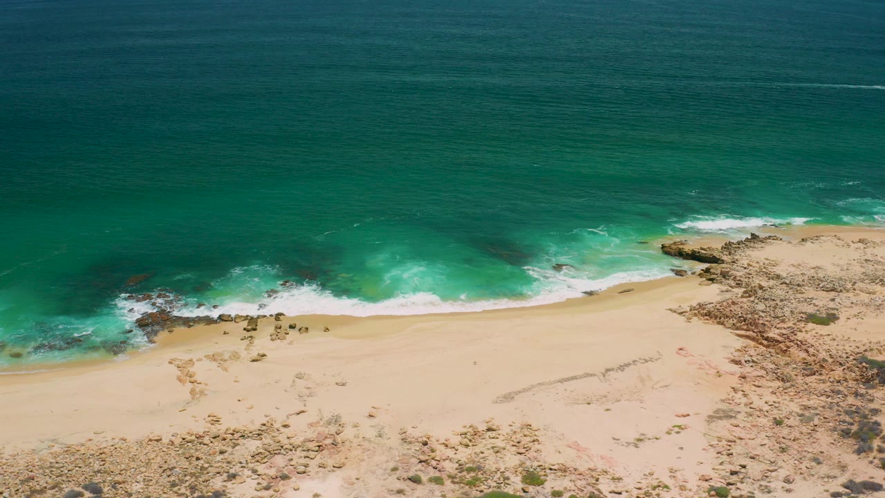 Aerial view over San Jose del Cabo coastline with teal ocean, white surf, pale sand, rocky shoreline patches, and no visible people.