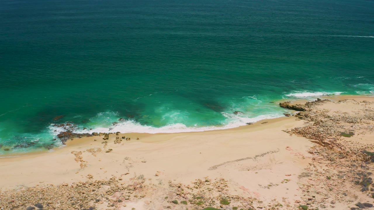 High aerial frame in San Jose del Cabo showing green-blue water, a thin surf line, broad sand, scattered rocks, and no visible people.