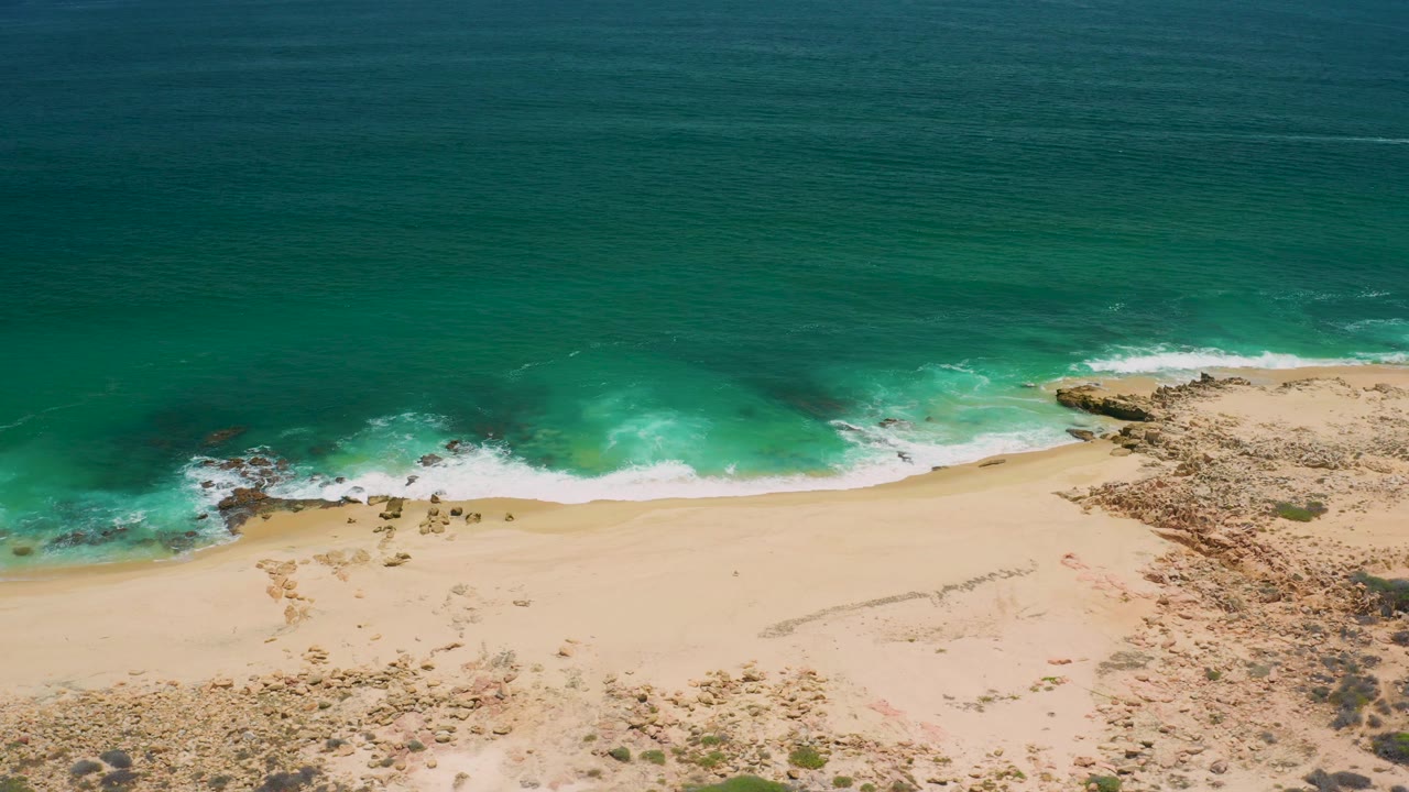 San Jose del Cabo aerial coastline with turquoise water across the top, white waves at center, empty sand below, rocky coast to the right, and no visible people.